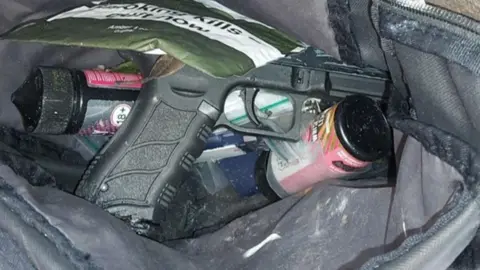 West Midlands Police A gun near other items. The gun is black and rests in a bag on top of a can and a small plastic bag with another bag with the words "smoking kills" resting on top of it.