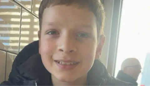 Family picture Aaron Anderson smiling while sitting inside a restaurant, which has white tiles. In the background is a man sitting in front of a large window who is wearing glasses and a black jacket.