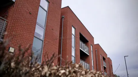 A three storey, red brick block of flats with balconies enclosed by glass and steel barriers. A slate grey sky. 