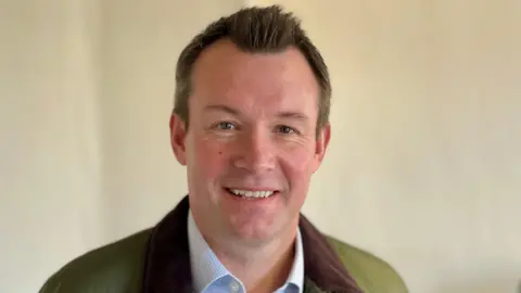 A man with brown hair is smiling at the camera. He is wearing a white and blue checked shirt with a green jacket that has a brown collar