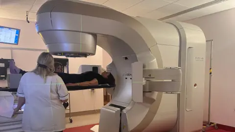 Trish Prosser Trish lying on a table as she is being scanned in a large silver hospitan scanner. There are two healthcare professionals in white uniforms standing next to her.
