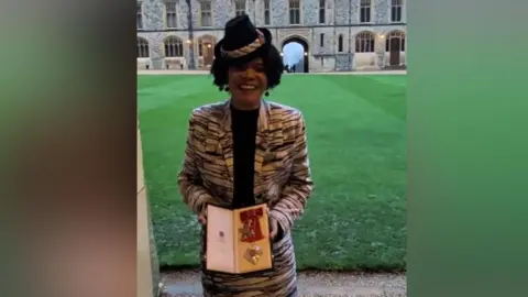 Chi Onwurah A slightly blurry photo of Dame Chi holding her damehood medal in the grounds of Windsor Castle. She is wearing the same black and white outfit and has a big smile on her face. The medal has a red bit of material attached to it.