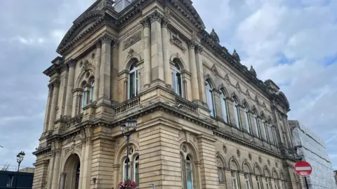 BBC Huddersfield Town Hall, which is a Grade II-listed building, with clouds gathering overhead. The back right hand side of the building has building work taking place, with scaffolding in place.