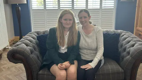 Summer is sitting next to her mother Karen on the couch in the family's living room. Summer is wearing her current school uniform which comprises of a black skirt, a white shirt and a green blazer.