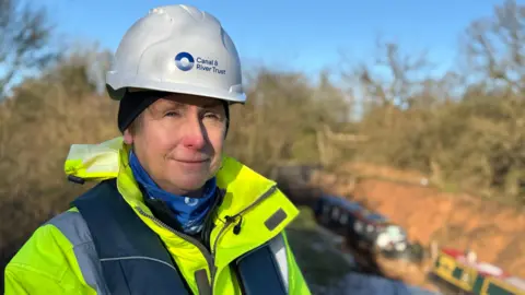 A woman wearing a white helmet with blue writing that says "canal and river trust". she is wearing a yellow hi vis jacket and is standing in front of the breach hole where you can see the two boats at the bottom of the hole blurred in the background