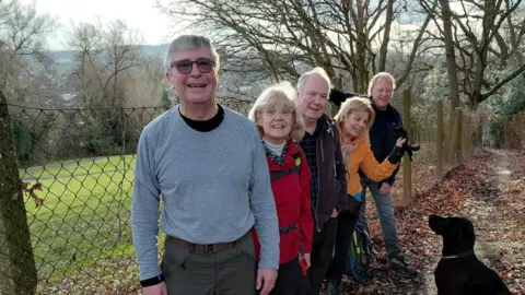 A group of walkers, they are lined up in a row on a country path with a black dog.