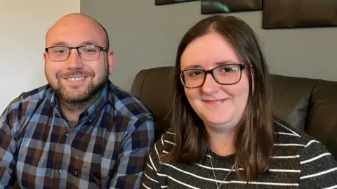 A balding man with a beard and glasses sitting on a sofa at home next to a woman with long brown hair and glasses 