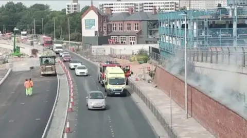 Cars and lorries drive along a dual carriageway. A fire engine and a yellow emergency service vehicle are parked in the righthand lane. A cloud of smoke can be seen on a grassed area. A blue multi-storey car park is visible in the background.