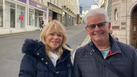Carol and Barry Shephard are standing in a street and smiling at the camera. On the left, Caroll has with blonde hair and is wearing a fluffy-hooded, blue coat. On the right is Barry and he has white hair and tinted spectacles, and he is wearing a blue jacket.