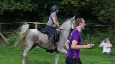 A man in a purple T-shirt runs through a field. Behind him, a woman rides a horse which also appears to be galloping. A spectator holding a phone up to take a picture can be seen in the background. 