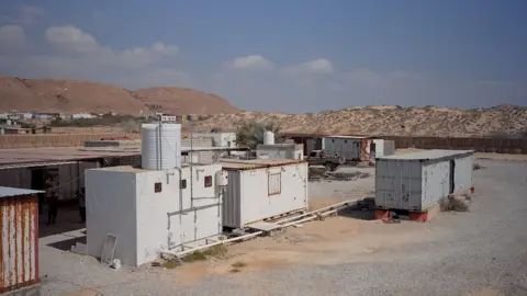 One white shipping container and two grey ones are seen on a gravel area with sandy hills behind them. A small white building with a water tank on it is next to one of them.