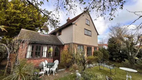Martin Giles/BBC A historic cottage, with a brick lower storey and rendered upper storeys. In the foreground is a garden featuring wrought iron garden furniture.