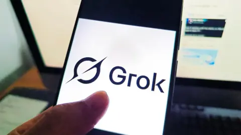 Getty Images A phone screen that displays the logo of Grok against a white background. There is a computer screen in the background also displaying Grok.