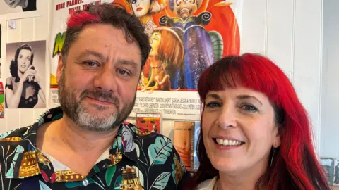 Leif and Sam looking at the lens, Sam has red and black hair and Leif has dark hair with a red highlight. Posters of old media can be seen in the background.