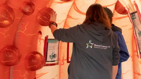 Bowel Cancer UK Two people with their backs to camera, one wearing a grey hoodie with the Bowel Cancer UK logo on the back, walking through a large, plastic pop-up tunnel simulating the inside of the bowel with large red round polyps