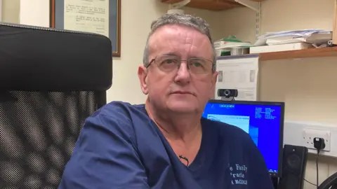 Dr. Alan McCulo sat on an office chair in his GP surgery. He wears a dark scrub with his surgery name on it. He has brown hair and wires wears rimd glasses. His expression is neutral. 