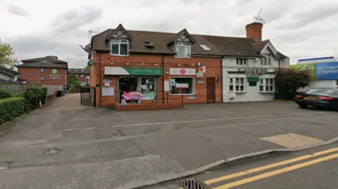 Google A building in the middle of the photo has the words post office in white on a green backing and to the right of that is a yellow and red post office logo. Part of a road is visible in the foreground.