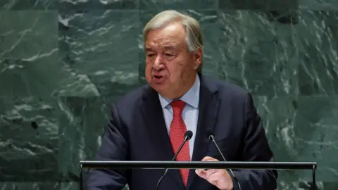 Reuters UN Secretary-General Antonio Guterres delivers a speech at the United Nations in New York. He stands at a lectern in front of the striking green marbled background and wears a red tie, a dark jacket and a blue shirt