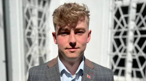 Simon Dedman/BBC Nathan Robins stands in front of a white building on a sunny day. He has blond hair which hangs over his forehead. He wears a grey suit jacket with a light blue shirt underneath.