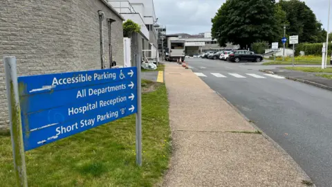 A blue sign at Guernsey's Princess Elizabeth Hospital, which points towards accessible parking, all departments, hospital reception and short stay parking. In the background is the entrance to the building, which is made of grey ribbed concrete. 