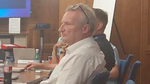 LDRS David Higgs sits in a health board meeting in a wood-panelled room. He has receding white hair and wears a white shirt and glasses pushed back over his head.