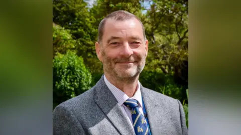 Simon Ring is smiling at the camera. He's wearing a grey, woollen suit jacket over a red and white checked shirt and a blue and gold tie.