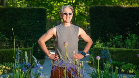 Alan Cumming standing in a garden with a pond behind him. He is wearing white sunglesses and a grey cut off T-shirt. he is standing smiling, with his hands  on his hips