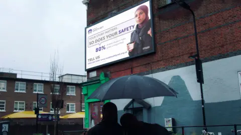 Muslim Women's Network UK A billboard advert for the helpline is positioned on a wall at the end of the street. Mariah and her double are stood beneath it looking up at it from under an umbrella. Part of the wall below the advert is painted light and dark blue.
