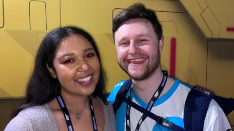 A woman with long dark hair wearing a grey jumper stands next to a man in a blue and white gaming jersey, both smiling at the camera. They are wearing DreamHack lanyards and standing in front of a large yellow and red display wall. The man is carrying a dark backpack over his gaming shirt.
