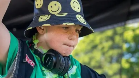 Efan Electra, pictured in a black bucket hat with yellow smiley faces on it. He wears headphones around his neck and purses his lips together. 