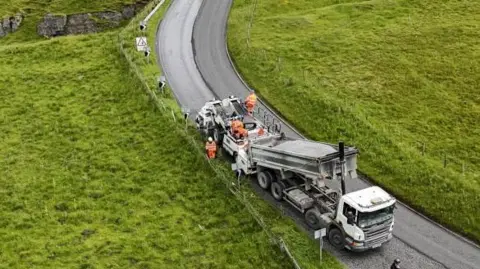 A lorry lowers chippings on to a road while workman sit on the back of the vehicle. The scene - a rural road with sweeping bend and green fields on either side - is pictured from above. The lorry has a white cab, in the foreground, and the trailer behind is hydraulic and tilted to about 25 degrees to allow the grey coloured contents to pour into other heavy equipment behind.