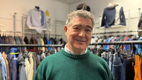 A middle aged man standing in front of a selection of clothing racks. He is wearing a green jumper and is smiling. 