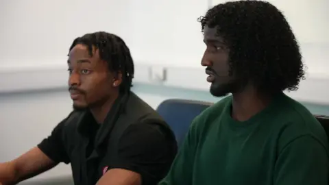 Tosin, 20, and Adnan, 18, sit at the table in an office room taking part in the discussion. Tosin, wearing a black sleeveless jacket, looks straight ahead, listening, while Adnan, in a dark green top, is speaking.