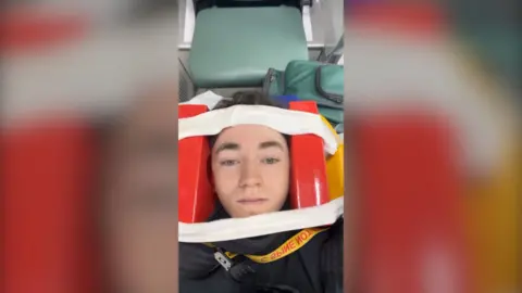 Supplied A selfie of Taylor in the ambulance after a crash caused by e-bike riders. He has red padded blocks either side of his head and pale material across the top and bottom of his face and wrapping around the back of the blocks. He is looking at the camera.