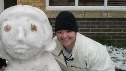 Ezzy Pearson A young woman wearing a black beanie and a big white coat smiles as she stands beside a snow woman in front of a building, potentially a classroom. There is snow on the grass behind her.