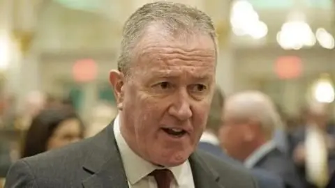 PA Conor Murphy, in a grey suit, white shirt and tie, appears to be animated or mid sentence. A woman and a man appear on either side of him, but in the background.