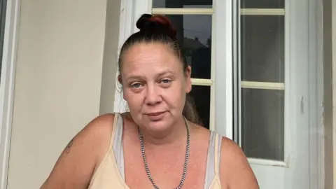 Head and shoulders image of Laura Smith who has dark hair tied in a bun outside a house in front of a white-framed glass door, wearing a light-coloured vest top and chain necklace. 
