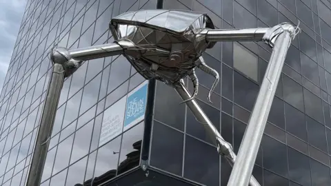 A shiny insect-like metal structure on three cylindrical legs stands in front of a tall glass-fronted building. There are two winding tentacles coming out the back of the statue.