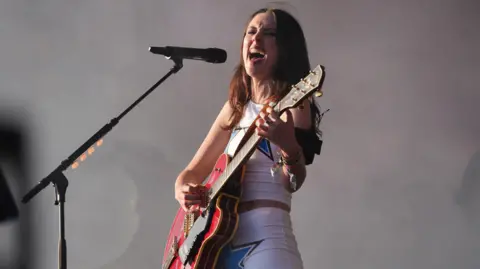 PA Media A woman with long dark hair stands on stage holding a red guitar and singing into a microphone on a stand. She is wearing a white crop top with blue stars on it and hot pants.