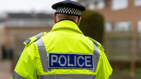 A man in a fluorescent yellow police jacket, walking away from camera. His back and head can be seen and he is wearing a black and white police hat. A blurred block of flats can be seen in the background. The word police is written across the back of his jacket.