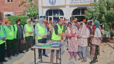 Paul Uppal A group of Sikh men in hi-vis vests and turbans standing near a large group of people - the people at the front are women in headscarves. A man hands over a red package to one of the women.