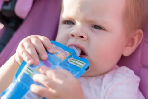 A young toddler holds a blue-coloured food pouch to its lips and is eating from it while sitting in a pram