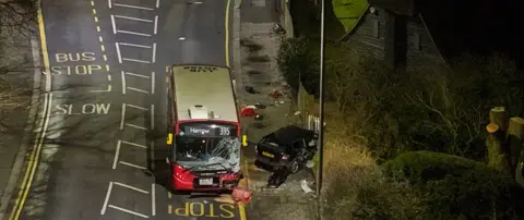 A 395 single decker bus with a smashed window and a black car which has crashed into a hedge 