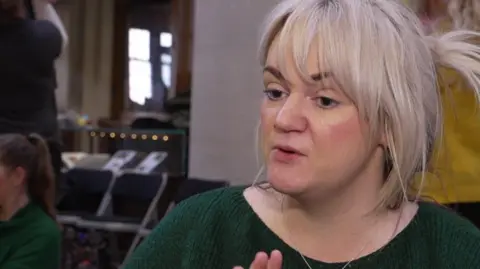 Becca with long blond hair in a pony tail wearing a dark green jumper at an event inside Liverpool Central Library. 