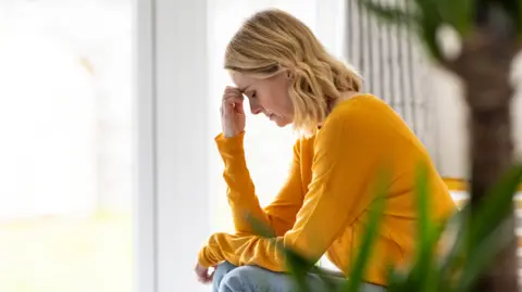 A woman with blonde, shoulder-length hair, sits facing to the left of the frame. She wears an orange top and light blue jeans. She has her forehead resting on the fingers of her right hand as her left hand lies in her lap. In the foreground is the trunk of a plant with some green leaves blurred in the foreground.