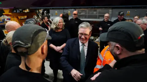 Reuters Sir Keir Starmer smiling as he meets workers at the Hitachi factory. He is wearing glasses, a navy suit, white shirt and a tie. Two workers in the foreground are facing away from the camera. They are wearing dark tops and green baseball caps.