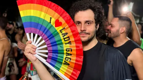 A man holds a rainbow fan which says I Love Lady Gaga