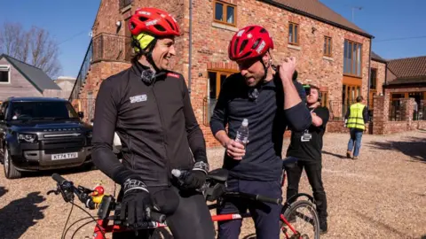 BBC/PA Greg James with brown curly hair smiling to his right in a red helmet and wearing a mic and black zip up jacket and leggings with gloves on with one hand on a tandem he is sitting on and the other holding a water bottle. Next to him is Prince William smiling and wearing a blue half-zipped up fleece and blue joggers, holding a bottle of water and looking towards the floor with a red helmet on. Behind them is the back of a pub with a brick exterior and long windows. A cameraman can be seen behind them and a person in high vis behind him.