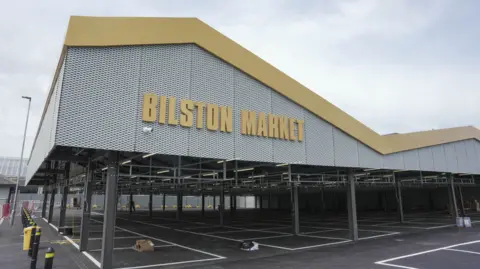 The market has a large grey and yellow roof section with pillars holding it up below. There are bollards at ground level. A yellow Bilston Market sign appear on the left side of the roof section.