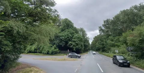 A T-road junction with a grassed island and trees and bushes surrounding the roads.